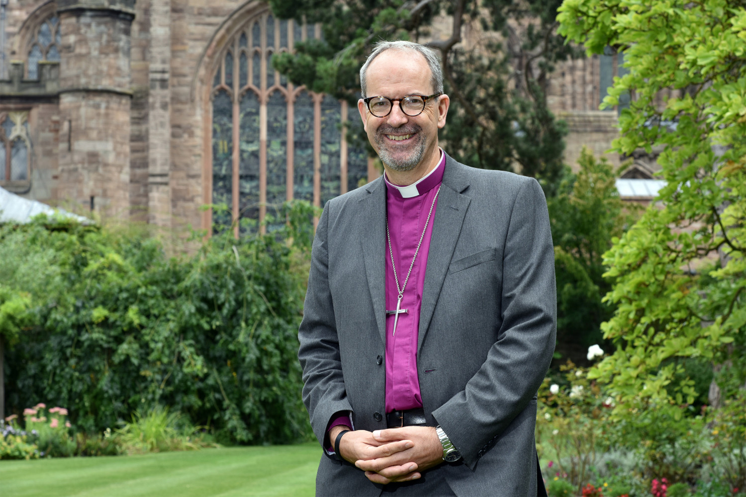 Male Bishop wearing purple shirt, crucifix necklace, grey suit and glasses stood on grass with Cathedral and tree in background