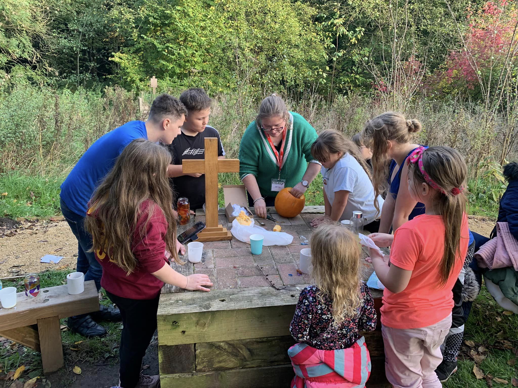 Group of children and adult outdoors preparing for a harvest celebration, carving pumpkins and praying together