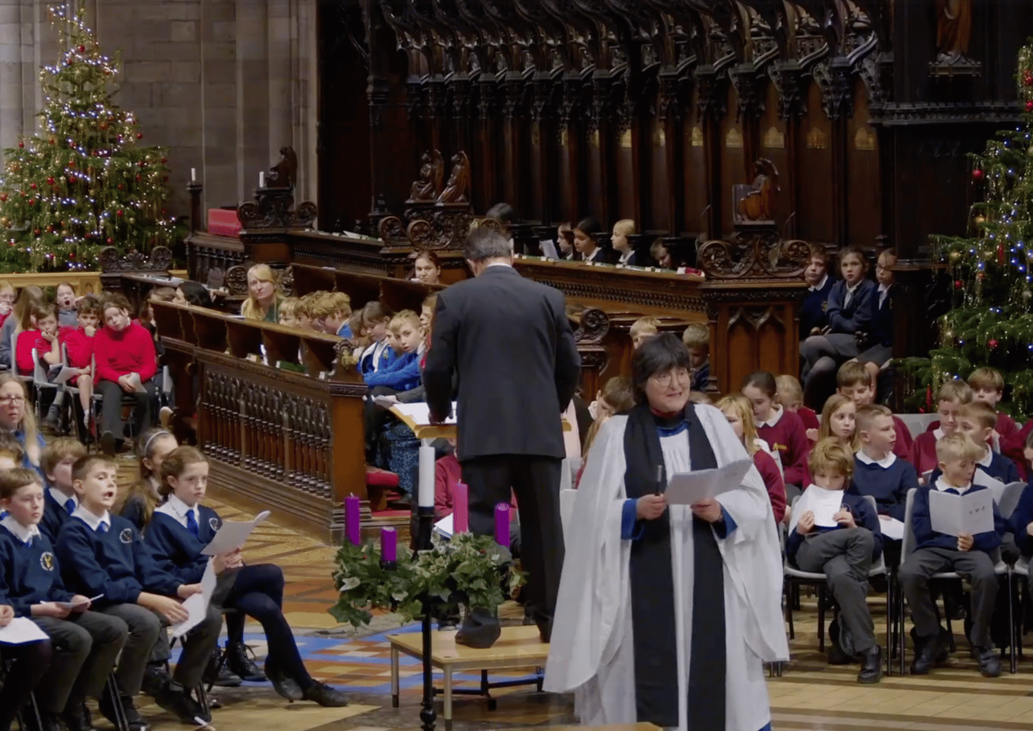 Hereford Cathedral with Dean Sarah stood reading at front of the altar, view of Cathedral Choir stalls filled with school children seated in groups, wearing different school uniforms