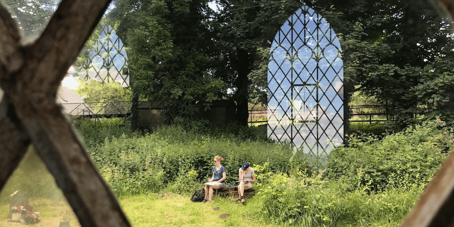 view looking from inside a church to outside, capturing the diamond shape of the window.  In the foreground two women sat on a stone bench sketching with trees in the background.