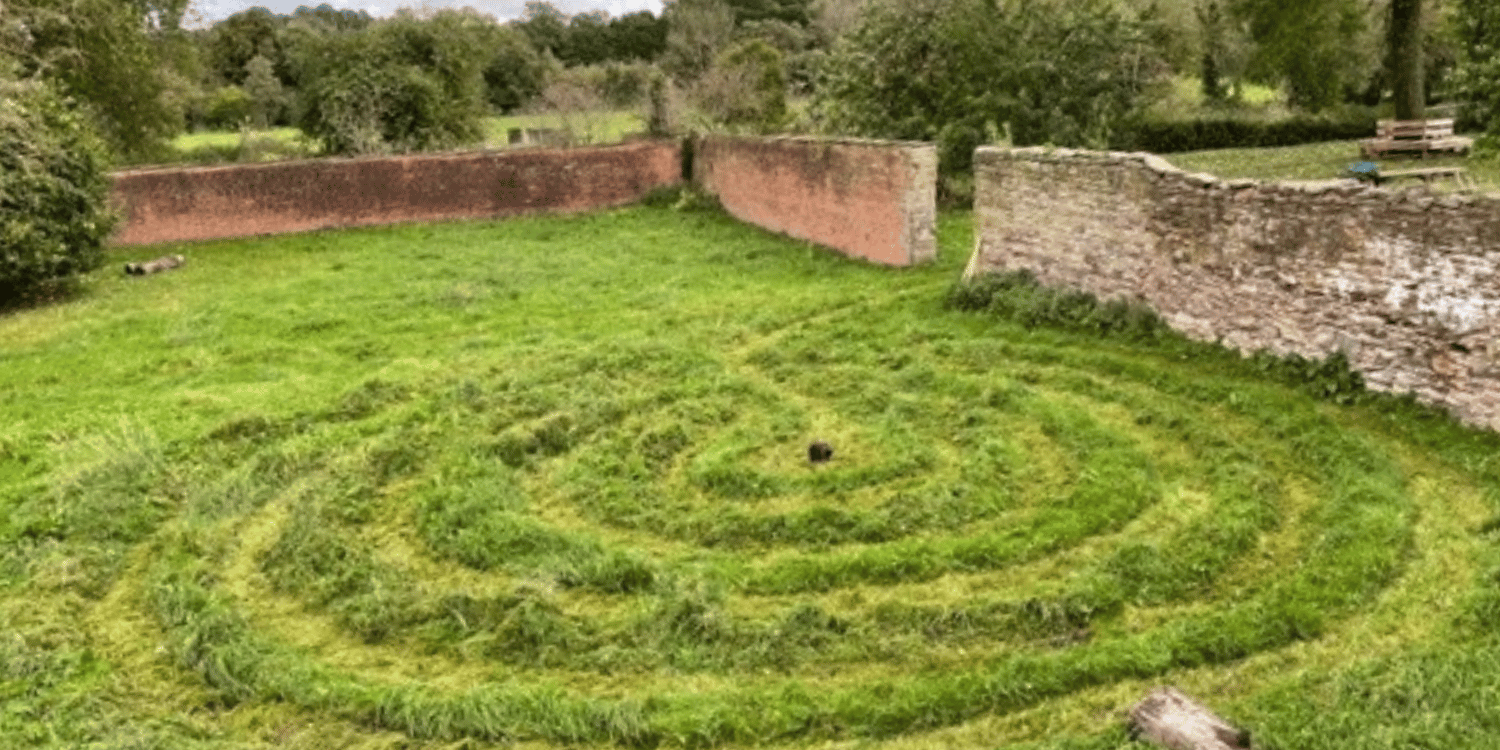 Image of garden with grass mown into a labyrinth surrounded by red brick wall and trees in the background