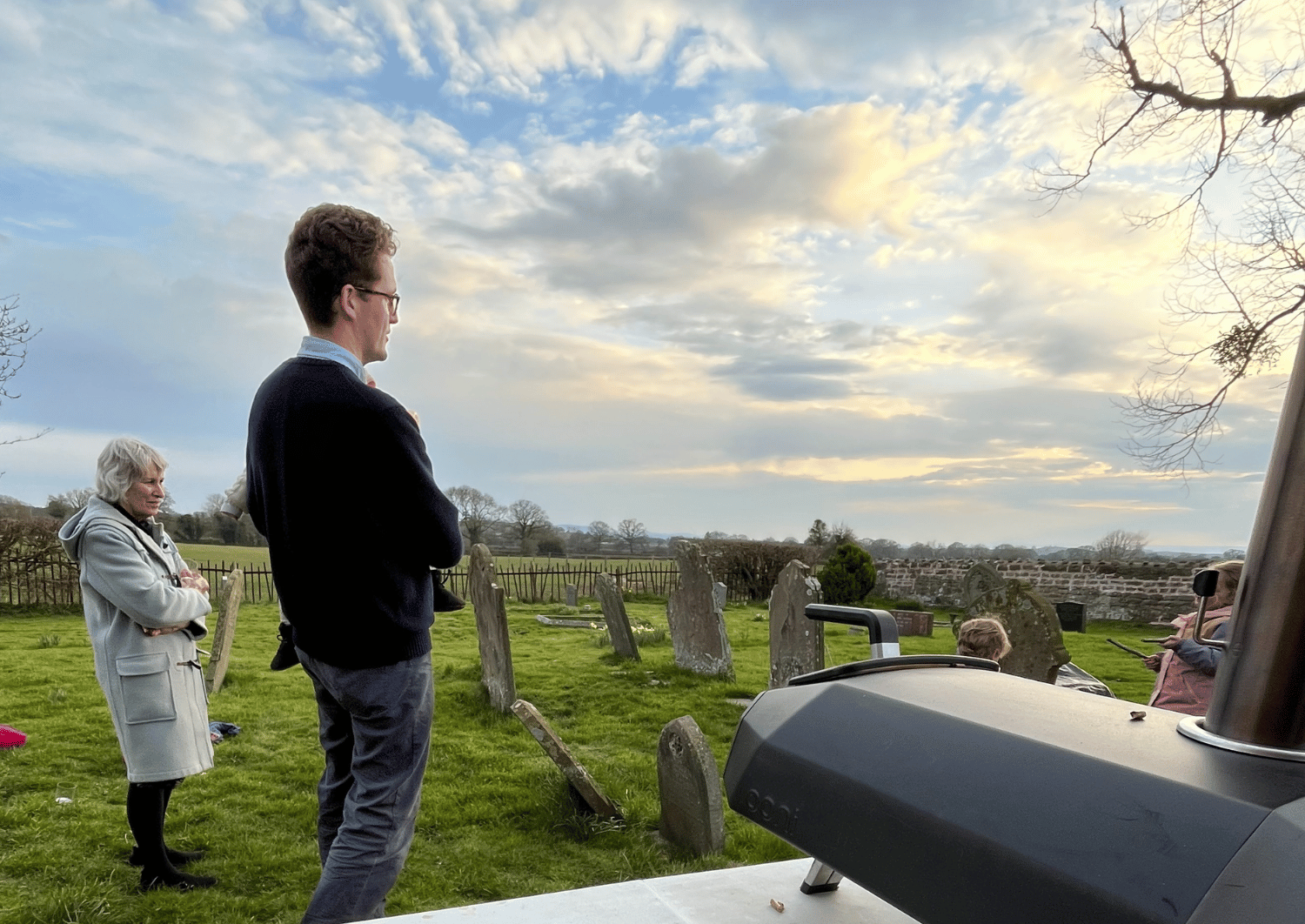 Man stood in churchyard holding a young child with a wintry blue cloudy sky and a pizza oven on the table in the foreground