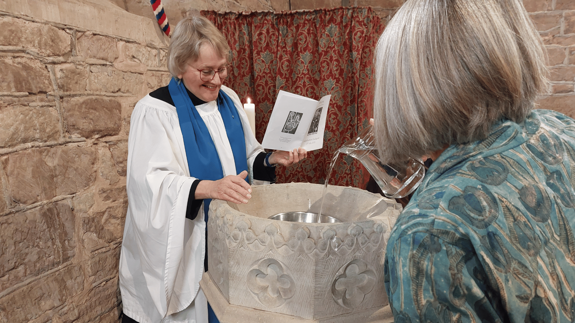 Diane Bates prepares and blesses holy water from font whilst woman pours water into font from glass jug