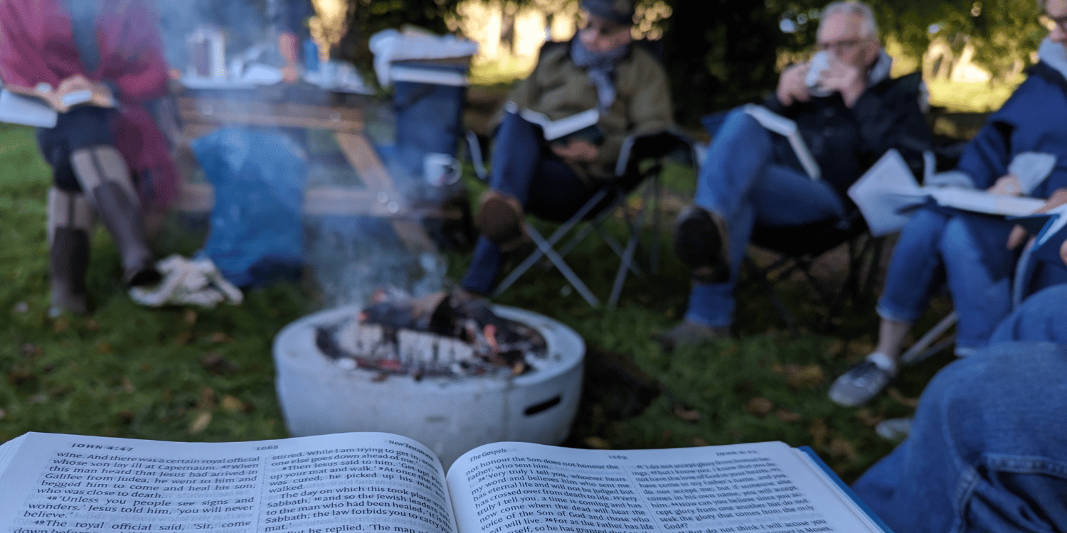 Bible open in front of a fire with soft focus people sat in background in deck chairs drinking coffee