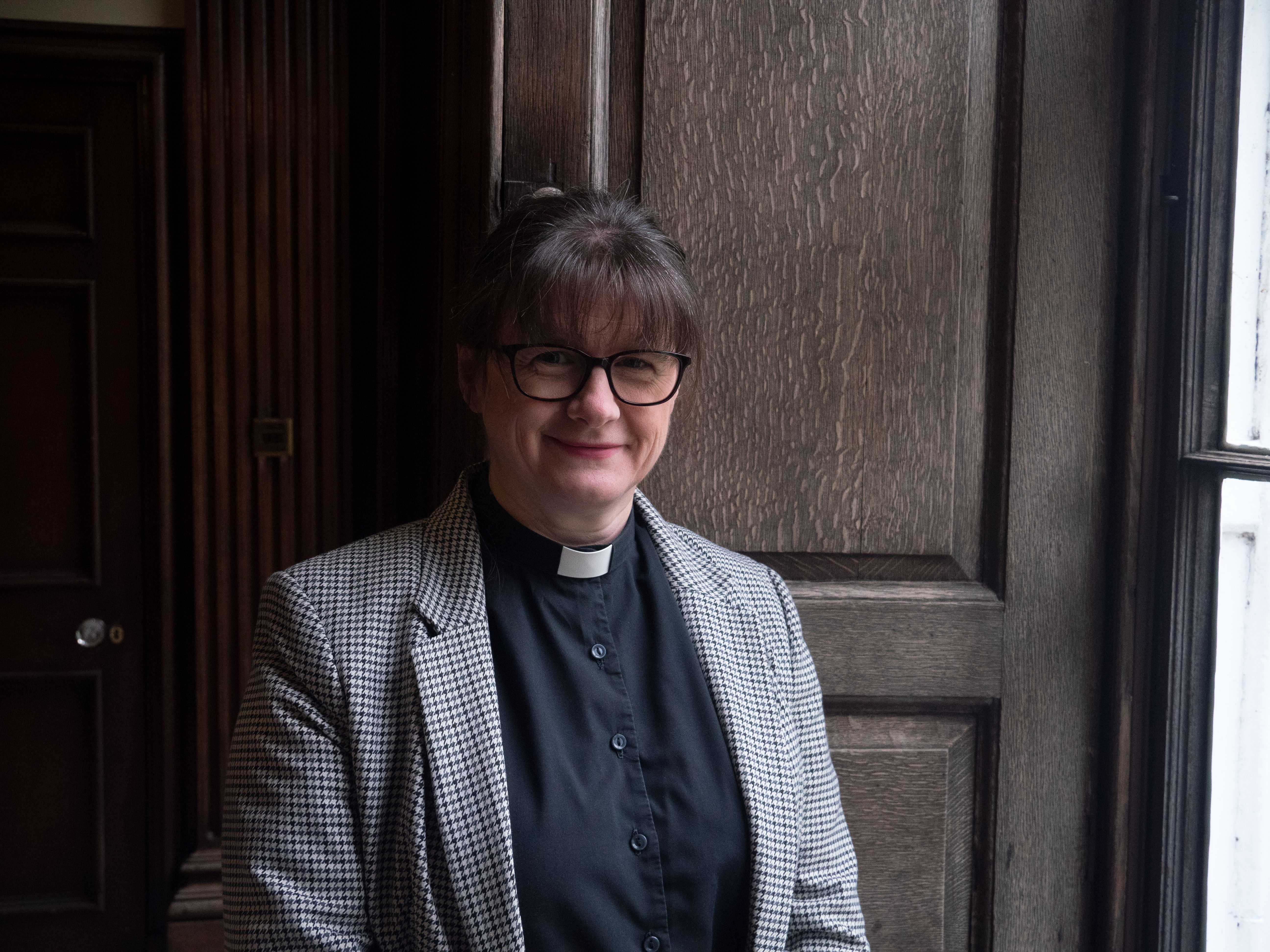 Archdeacon Fiona stood in a window wearing a houndstooth black and white check jacket with her dog collar, a pair of glasses and smiling to the camera