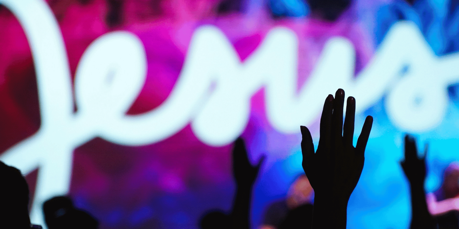 Image of hands being held up in praise with neon Jesus sign in background