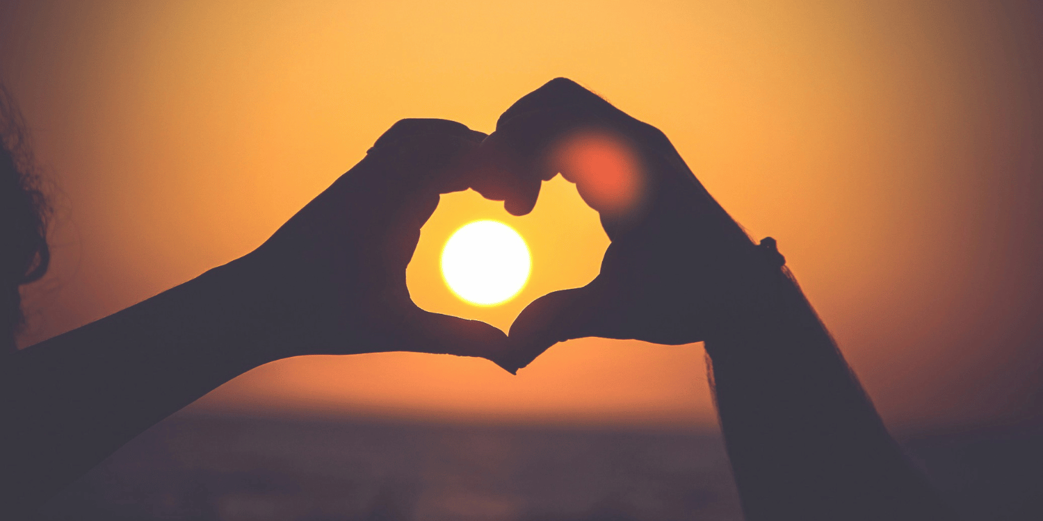 Hands making a heart shape around a setting sun in the background