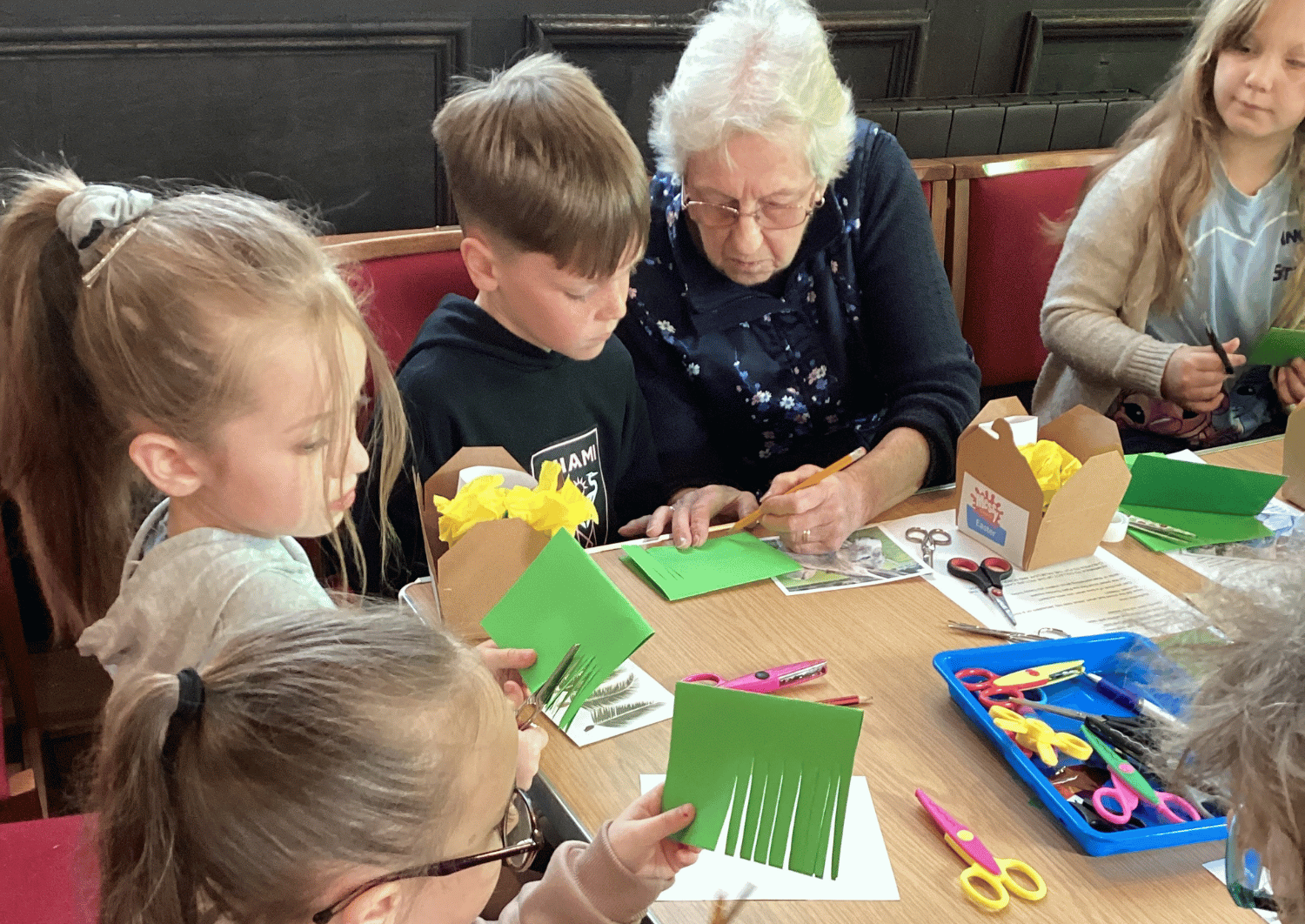Young children sat around a table with an older lady making craft items, one child is using a pair of scissors to cut paper and the old lady is drawing something for the young boy